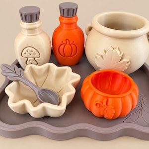 May include: A set of silicone kitchenware including a beige bottle with a mushroom icon, an orange bottle with a pumpkin icon, a beige cauldron with a leaf, a beige leaf-shaped bowl with a grey spoon, and an orange pumpkin bowl on a grey tray.