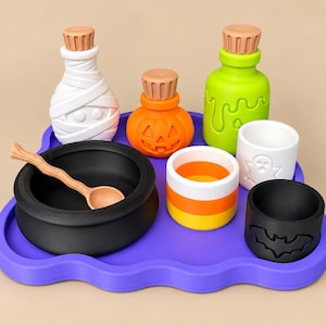 May include: A purple tray holding a black cauldron with a tan spoon, a white mummy bottle, an orange pumpkin jar, a lime green slime bottle, a candy corn cup, a white ghost cup, and a black bat cup, all featuring cork-style stoppers.