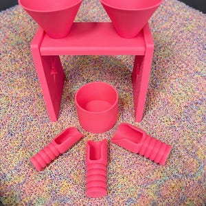 May include: A bright pink sensory play set consisting of a stand with two attached funnels, a small cylindrical container, and three handheld scoops with ribbed handles, all arranged on a bed of multicoloured, textured sensory filler material.