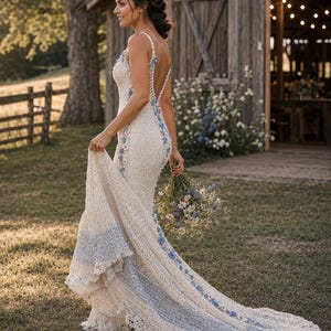 May include: A floor-length cream-colored crochet dress featuring a deep back, thin straps, and a flared hem. The garment is accented with blue floral embroidery along the straps, back, and a wide blue band detail near the bottom of the train.