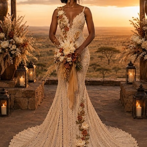 May include: A floor-length beige crochet gown featuring a deep V-neckline, thin straps, and a fitted bodice that flares into a wide hem. The dress is adorned with intricate floral appliqués in shades of cream, rust, and brown along the shoulder and skirt.
