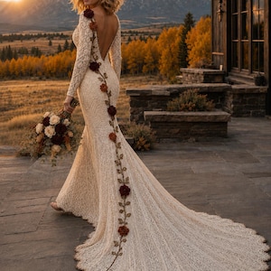 May include: A floor-length cream lace dress with long sleeves and a deep open back. A vine of dark red and brown fabric flowers runs from the shoulder down the center of the back and along the train. The dress features a scalloped hemline.