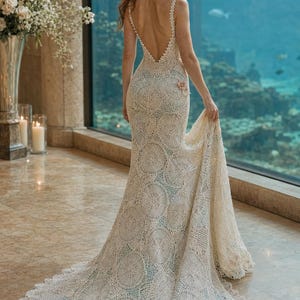 May include: A floor-length, cream-colored crochet gown featuring a deep V-back, thin straps, and a circular pattern throughout the fabric. The dress has a long train and is accented with small, pale pink floral embellishments at the hip and hem.