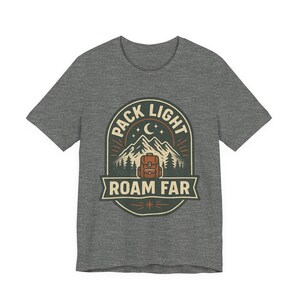 May include: A heather gray short-sleeve t-shirt featuring a graphic print of a mountain range, a backpack, and a crescent moon. The text "PACK LIGHT" is arched above the mountains, and "ROAM FAR" is printed on a banner below the backpack.