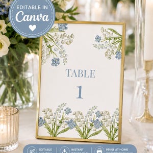 Op de afbeelding: Een tafelnummerkaart met een gouden lijst, versierd met illustraties van blauwe en witte bloemen. De tekst "TABLE 1" is blauw. De kaart is ontworpen om te worden bewerkt in Canva. Ideaal voor bruiloften en evenementen.