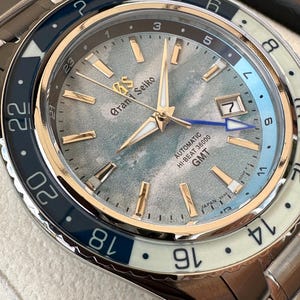 May include: A Grand Seiko automatic watch with a textured light blue and grey dial, gold-tone hands and indices, and a date window at the three o'clock position. The bezel features a two-tone blue and white GMT scale, and the watch includes a blue GMT hand.