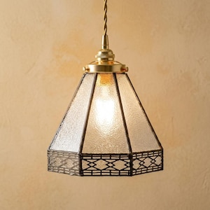 May include: A pendant light featuring a hexagonal, textured glass shade with a dark metal frame. The base of the shade has a geometric lattice pattern. It is suspended by a twisted cord from a polished brass fixture with a cylindrical top.