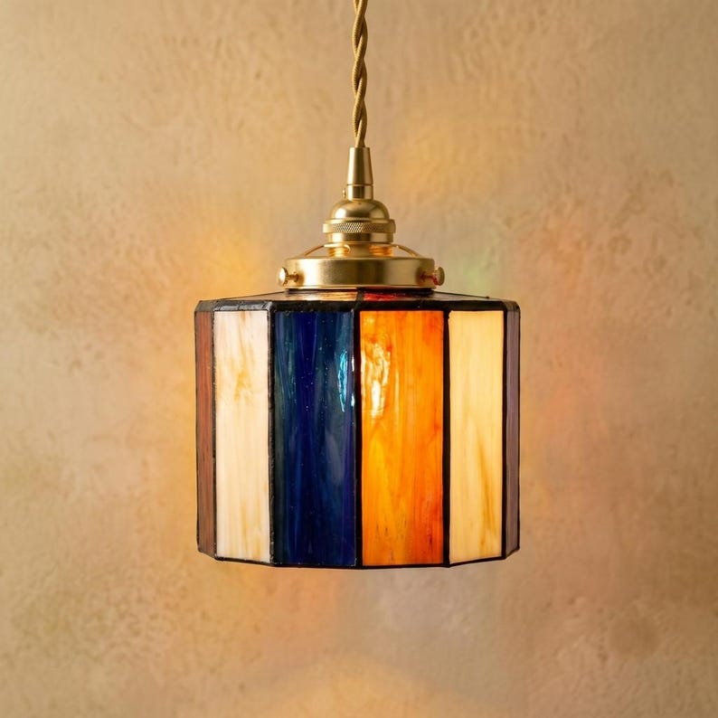 K&ouml;nnte beinhalten: Eine sechseckige Buntglas-H&auml;ngeleuchte mit abwechselnden vertikalen Paneelen aus cremefarbenem, tiefblauem und leuchtend orangefarbenem Glas. Die Lampe h&auml;ngt an einem gedrehten Kabel und hat eine Kappe sowie Beschl&auml;ge aus poliertem Messing.