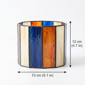 K&ouml;nnte beinhalten: Ein zylindrischer Kerzenhalter aus Buntglas mit vertikalen Paneelen in Creme, Blau und Orange, die durch dunkle Metalll&ouml;tstellen verbunden sind. Das Objekt ist 12 cm hoch und 13 cm breit, wie durch die sichtbaren Ma&szlig;angaben dargestellt.