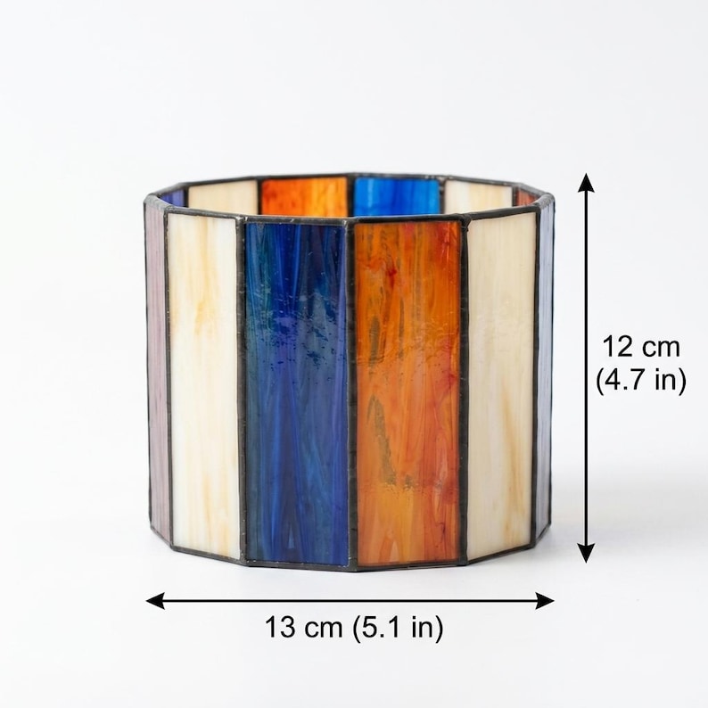 K&ouml;nnte beinhalten: Ein zylindrischer Kerzenhalter aus Buntglas mit vertikalen Paneelen in Creme, Blau und Orange, die durch dunkle Metalll&ouml;tstellen verbunden sind. Das Objekt ist 12 cm hoch und 13 cm breit, wie durch die sichtbaren Ma&szlig;angaben dargestellt.