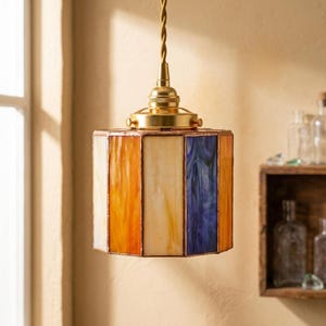 K&ouml;nnte beinhalten: Eine zylindrische H&auml;ngeleuchte mit Buntglas-Paneelen in abwechselnden Farbt&ouml;nen von Orange, Creme und Blau. Die Glassegmente sind durch d&uuml;nne Metalllinien verbunden, und die Leuchte ist an einem gedrehten Kabel an einer Messinghalterung befestigt.