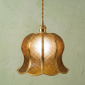 May include: A bell-shaped pendant light featuring amber-colored textured glass panels with a repeating floral pattern. The fixture is held by a metallic gold-toned frame and a matching twisted cord, creating a warm, glowing illumination effect.
