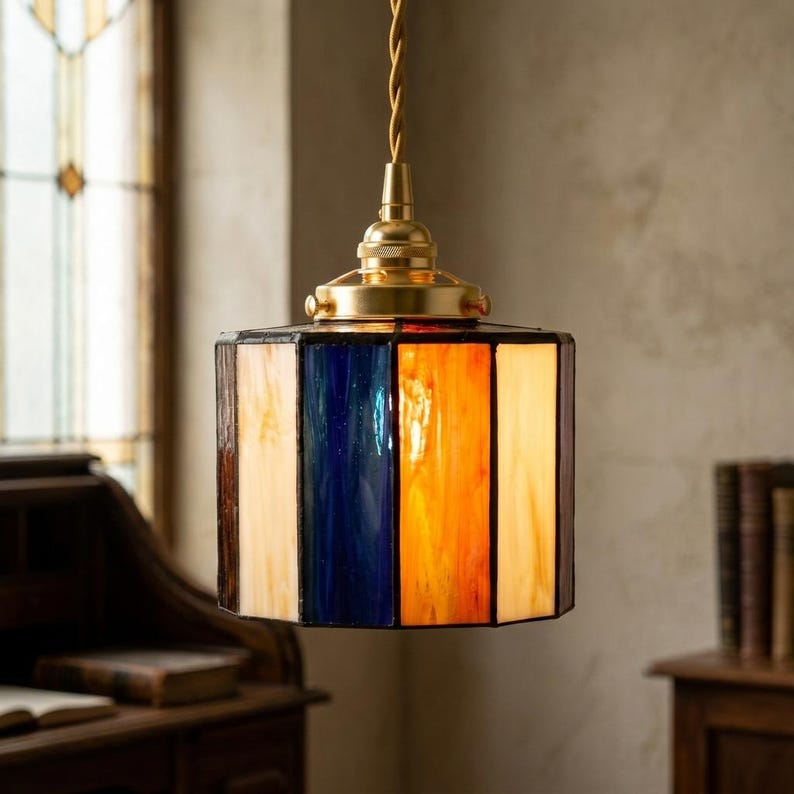 K&ouml;nnte beinhalten: Eine Pendelleuchte mit einem zylindrischen Lampenschirm aus Buntglas, der aus abwechselnden vertikalen Paneelen in tiefem Blau, warmem Orange und Cremefarben besteht. Der Schirm ist an einer messingfarbenen Metallkappe und einem gedrehten Kabel befestigt.