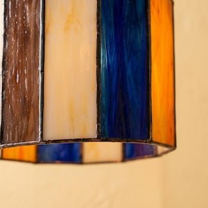 K&ouml;nnte beinhalten: Ein zylindrischer Lampenschirm aus Buntglas mit vertikalen Paneelen aus braunem, cremefarbenem, tiefblauem und orangefarbenem Glas. Die Paneele sind durch dunkle Metalll&ouml;tstellen verbunden und bilden eine geometrische Struktur, die Licht durchl&auml;sst.