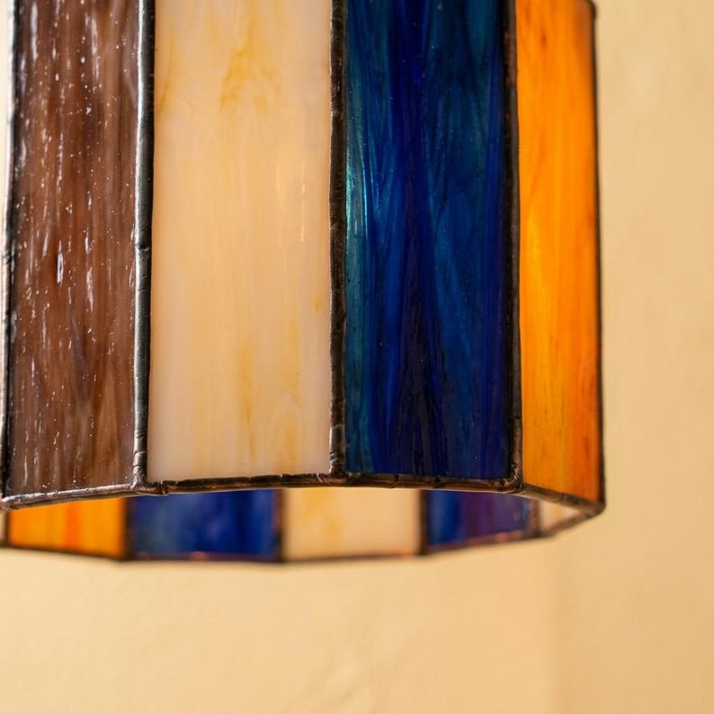 K&ouml;nnte beinhalten: Ein zylindrischer Lampenschirm aus Buntglas mit vertikalen Paneelen aus braunem, cremefarbenem, tiefblauem und orangefarbenem Glas. Die Paneele sind durch dunkle Metalll&ouml;tstellen verbunden und bilden eine geometrische Struktur, die Licht durchl&auml;sst.