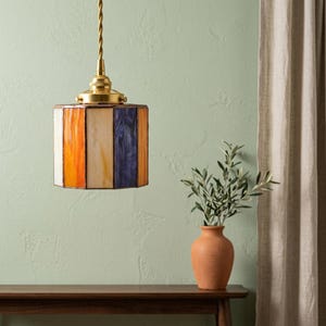 K&ouml;nnte beinhalten: Eine h&auml;ngende Pendelleuchte mit einem zylindrischen Schirm aus vertikalen Buntglaspaneelen in Orange-, Creme- und Violettt&ouml;nen. Die Lampe ist an einem gedrehten Kabel an einer Kappe und Befestigungselementen aus poliertem Messing aufgeh&auml;ngt.