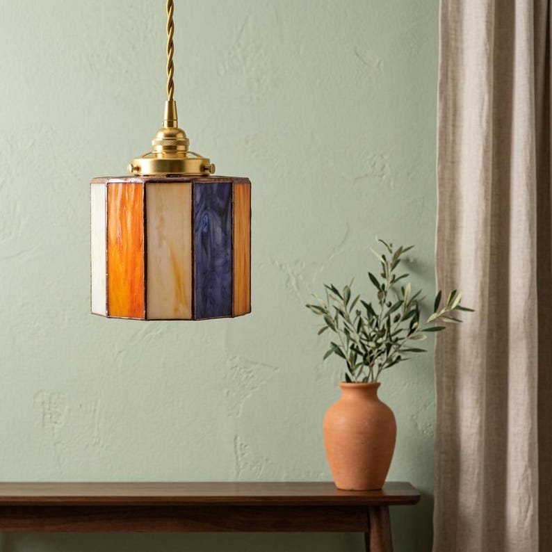 K&ouml;nnte beinhalten: Eine h&auml;ngende Pendelleuchte mit einem zylindrischen Schirm aus vertikalen Buntglaspaneelen in Orange-, Creme- und Violettt&ouml;nen. Die Lampe ist an einem gedrehten Kabel an einer Kappe und Befestigungselementen aus poliertem Messing aufgeh&auml;ngt.