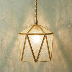 May include: A pendant light fixture featuring a geometric, multi-faceted shade constructed from textured, patterned glass panels held together by a gold-toned metal frame, suspended from a matching gold-toned cord and ceiling mount.