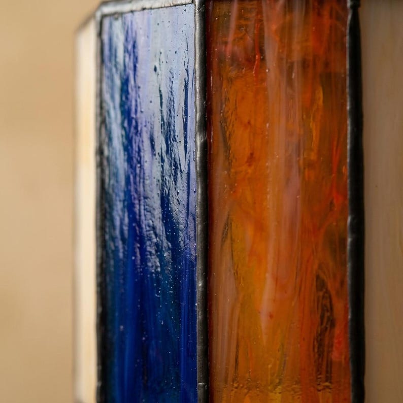 K&ouml;nnte beinhalten: Ein Buntglasfenster mit vertikalen rechteckigen Segmenten aus strukturiertem tiefblauem und leuchtend orangefarbenem Glas. Jedes Glasst&uuml;ck ist von dunklen, gel&ouml;teten Metalllinien umrahmt, die eine klare Gitterstruktur auf der Oberfl&auml;che bilden.