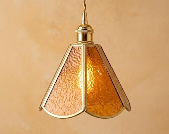 May include: A pendant light fixture featuring a conical shade made of textured amber-colored glass panels held together by a polished brass frame, suspended from a matching brass socket and a twisted gold-colored fabric cord.