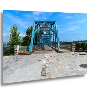 May include: A bright blue metal truss bridge with a wooden deck extends into the distance under a clear blue sky. The bridge features intricate steel framework, stone pillars, and white railings, set against a backdrop of green trees and foliage.