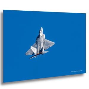 May include: A gray F-22 Raptor fighter jet in flight against a clear blue sky, viewed from a high angle. The aircraft displays a metallic finish, dual engine nozzles with orange exhaust, and the text Alan Locke Photography in the bottom corner.