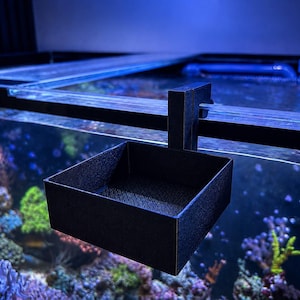Könnte beinhalten: Ein schwarzer, quadratischer Futterring für Aquarien mit strukturierter Oberfläche und einer Hakenbefestigung, die sicher über den Rand eines Glasaquariums gehängt werden kann, um schwimmendes Fischfutter an einer Stelle zu halten.