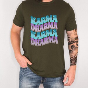 May include: An olive green short-sleeve t-shirt featuring a repeating wavy graphic print of the words KARMA and DHARMA in alternating shades of bright blue and light purple. The text is stacked in four rows with a distinct retro-inspired font style.