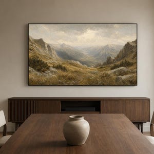 Puede incluir: Un cuadro de paisaje rectangular en un marco negro fino que representa un valle de montaña con hierbas de color marrón dorado, laderas rocosas y un cielo nublado. La obra está montada en una pared lisa sobre un aparador de madera.