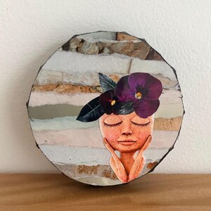 May include: A circular decorative plaque featuring a collage of a person with closed eyes and hands touching their cheeks. The person is adorned with dark purple pansy flowers on their head. The background consists of horizontal stone-like layers.