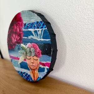 May include: A circular mixed media collage art piece featuring a stylized face with closed eyes and hands touching the cheeks. The artwork incorporates torn paper strips in shades of blue, a large dark pink flower, and a floral crown with green leaves.