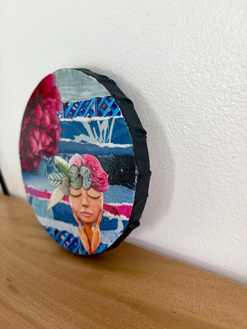 May include: A circular mixed media collage art piece featuring a stylized face with closed eyes and hands touching the cheeks. The artwork incorporates torn paper strips in shades of blue, a large dark pink flower, and a floral crown with green leaves.