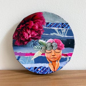 May include: A circular mixed media collage art piece featuring a large dark pink peony in the upper left corner. The background consists of horizontal strips of blue, white, and magenta paper. A central figure with closed eyes wears a floral crown.