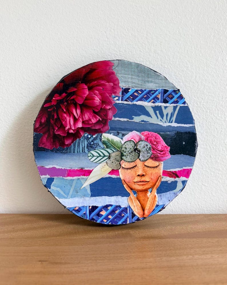May include: A circular mixed media collage art piece featuring a large dark pink peony in the upper left corner. The background consists of horizontal strips of blue, white, and magenta paper. A central figure with closed eyes wears a floral crown.