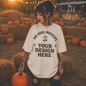May include: An off-white short-sleeve t-shirt featuring black text that reads INK ROSE MOCKUPS YOUR DESIGN HERE with a rose icon and star symbols. The shirt is worn by a person with visible arm tattoos, holding a large orange pumpkin.