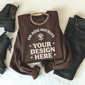 May include: A dark brown crewneck sweatshirt featuring a white graphic with a rose icon, four-pointed stars, and the text "INK ROSE MOCKUPS YOUR DESIGN HERE". The sweatshirt is displayed alongside a pair of black lace-up boots and black jeans.