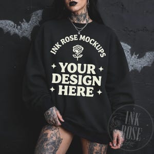 May include: A black crewneck sweatshirt featuring a cream-colored graphic print on the chest. The text reads INK ROSE MOCKUPS with a rose icon above the words YOUR DESIGN HERE, flanked by four-pointed star symbols on the left and right sides.