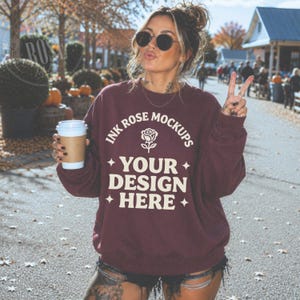 May include: A maroon long-sleeve crewneck sweatshirt featuring white text that reads INK ROSE MOCKUPS, a rose icon, and the text YOUR DESIGN HERE, accented with four-pointed stars. The sweatshirt is worn with dark denim shorts and sunglasses.