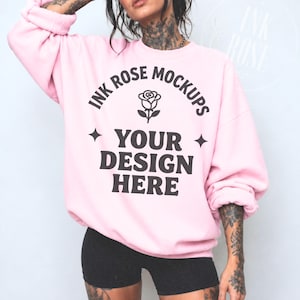 Op de afbeelding: Een lichtroze sweater met ronde hals en donkergrijze tekst met de tekst INK ROSE MOCKUPS, een roos-icoon en de woorden YOUR DESIGN HERE, geflankeerd door twee vierpuntige sterren. De sweater heeft lange mouwen en een relaxte pasvorm.