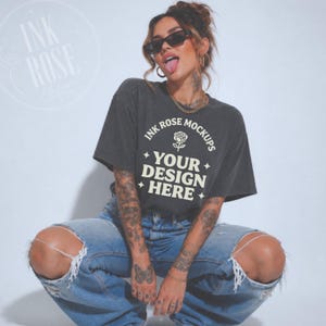 May include: A charcoal gray short-sleeve t-shirt featuring a white graphic print that reads INK ROSE MOCKUPS with a rose icon and the text YOUR DESIGN HERE surrounded by four-pointed stars. The shirt is paired with light blue distressed denim jeans.