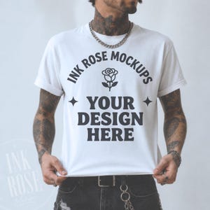 Może przedstawiać: Biały t-shirt z krótkim rękawem z czarnym napisem INK ROSE MOCKUPS, ikoną róży oraz napisem YOUR DESIGN HERE, otoczonymi dwiema czteroramiennymi gwiazdami. Koszulka jest noszona ze srebrnym łańcuszkiem i czarnymi dżinsami.
