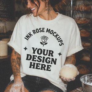 May include: An off-white short-sleeve t-shirt featuring black text that reads INK ROSE MOCKUPS, a rose icon, and the words YOUR DESIGN HERE, accented by two four-pointed star symbols. The shirt has a crew neckline and rolled sleeves.