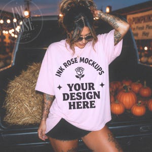 May include: A light pink short-sleeve t-shirt featuring black text that reads INK ROSE MOCKUPS, a rose icon, and YOUR DESIGN HERE, accented by two four-pointed stars. The shirt is displayed in a setting with hay and pumpkins in the background.
