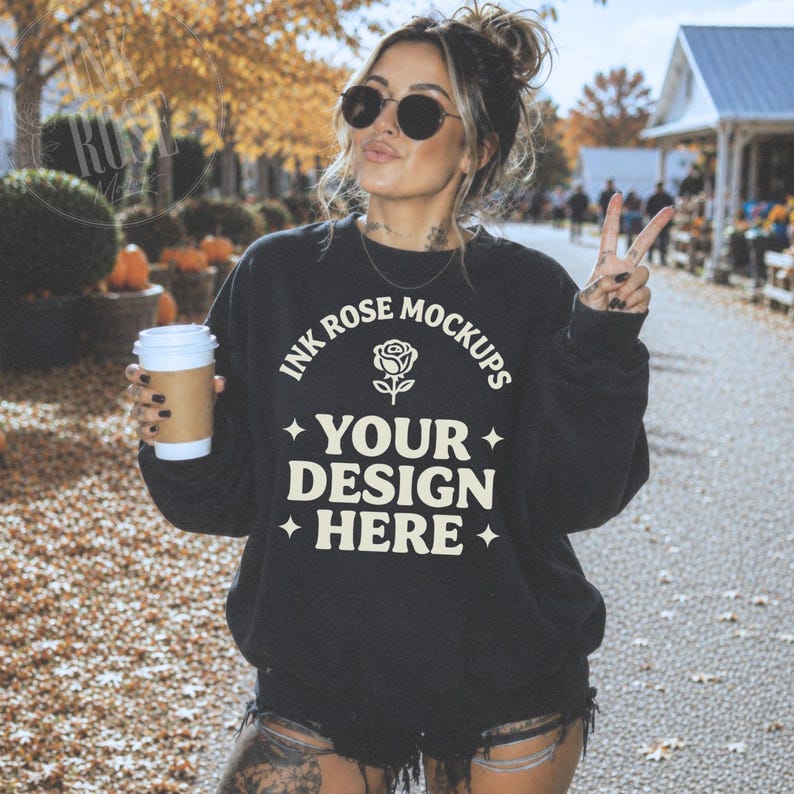 Tattooed woman wearing an oversized Gildan 18000 black sweatshirt while holding a coffee cup and flashing a peace sign at a fall market with pumpkins and autumn leaves.