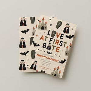 May include: Two rectangular birthday invitation cards with a cream background featuring a pattern of vampires, coffins, bats, and castles. The text reads: LOVE AT FIRST BITE, JOIN US TO CELEBRATE CHARLOTTE'S 1ST BIRTHDAY, SATURDAY, OCTOBER 14 AT 2 PM, THE ANDERSON HOME, 123 PUMPKIN LANE, AUSTIN, TEXAS, PLEASE RSVP BY OCTOBER 1ST, 512.555.0100.
