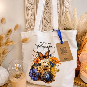 Puede incluir: Una bolsa de tela blanca con un estampado gráfico de una cámara rodeada de flores naranjas y azules y una mariposa monarca. El texto "AMANTES A LA fotografia" está impreso sobre la cámara. Una etiqueta marrón cuelga de una cinta azul.