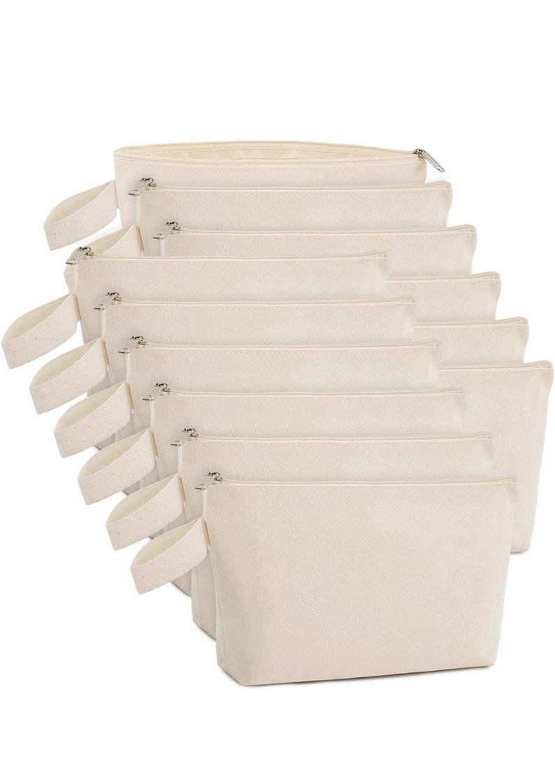 May include: A stack of eight rectangular off-white canvas pouches arranged in a cascading row. Each pouch features a top zipper closure with a silver-toned metal pull and a matching fabric wrist strap attached to the left side of the bag.