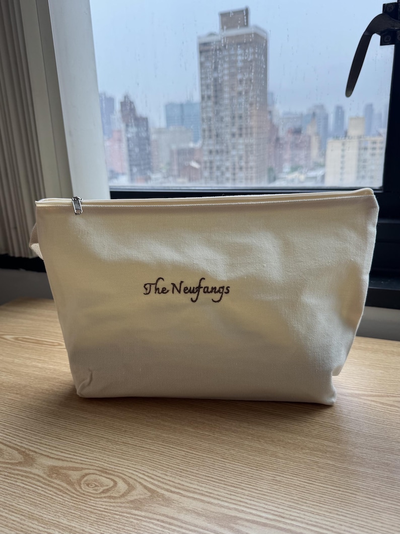 May include: A cream-colored canvas zippered pouch featuring the embroidered text The Neufangs in dark brown thread. The pouch has a rectangular base and a top zipper closure with a silver-toned metal pull, providing a simple storage solution.