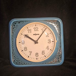 Könnte beinhalten: Eine quadratische Wanduhr mit hellblauem Rahmen und einem gesprenkelten, dunkelblau-braunen Glasurrand. Das Zifferblatt ist cremefarben mit schwarzen arabischen Ziffern von 1 bis 12, schwarzen Zeigern und dem in Schwarz gedruckten Markennamen PEWETA.