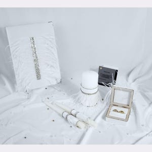 May include: A white wedding set featuring a rectangular guest book with a vertical rhinestone strip and white feathers, a pillar candle with a feathered base, two taper candles with rhinestone accents, and an open ring box with two gold rings.